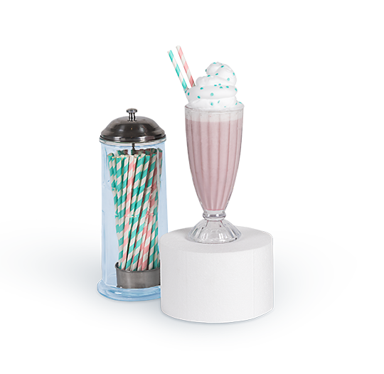 A glass jar filled with green-and-white striped paper straws sits beside a tall milkshake glass topped with whipped cream and two striped straws, both displayed on a round white pedestal”